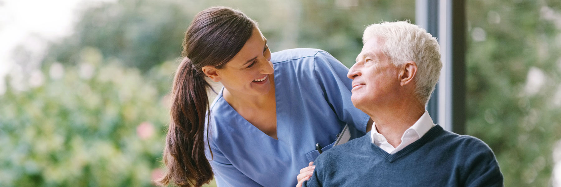 A smiling nurse rests her hands on an elderly patient’s shoulders. 