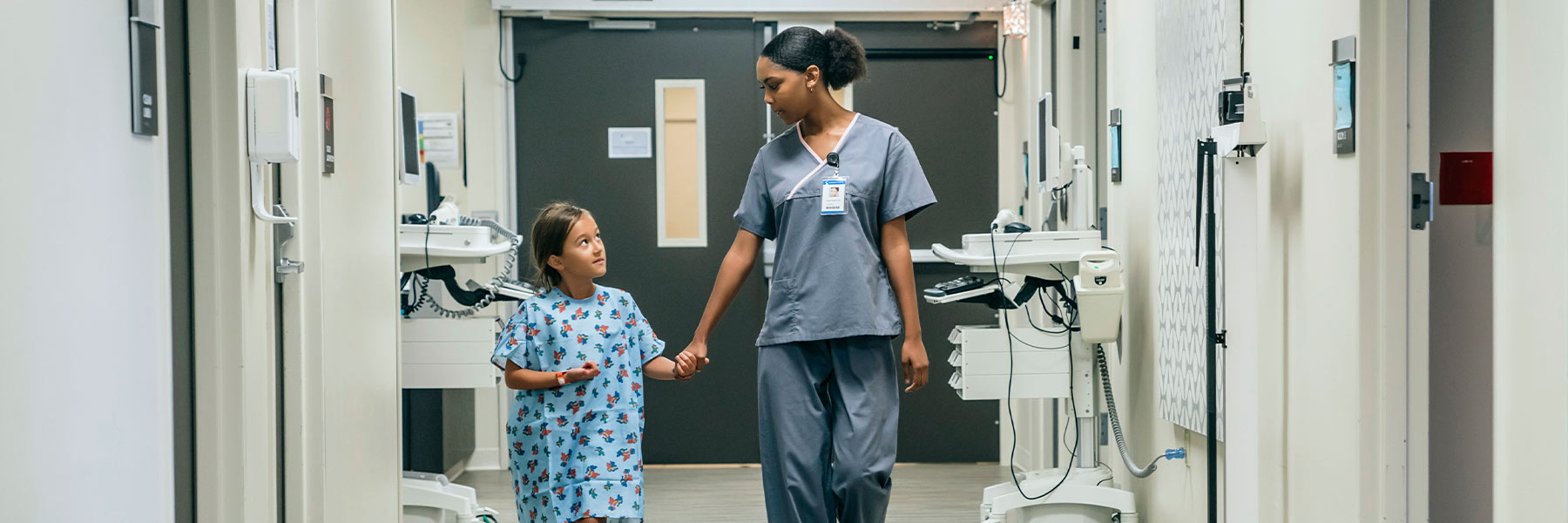 A nurse holds the hand of a small child as they walk through a hospital. 